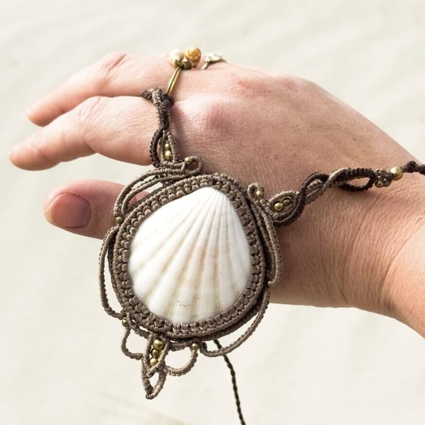 Handmade macrame necklace Of a white shell with light and brown accents and brass beads holding in one hand at the beach