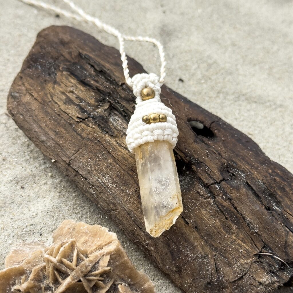 Handmade beige macrame necklace With tangerine quartz crystal and brass beads lying on a piece of driftwood on the beach
