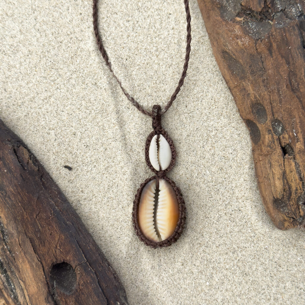 Handmade macrame necklace with two cowrie shells on top of each other. Displayed on the beach between driftwood.
